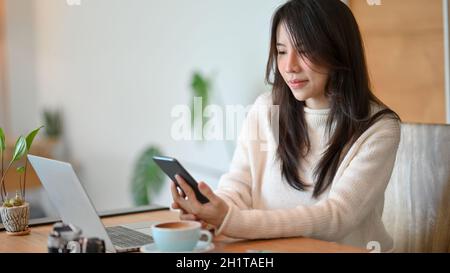A lovely Asian young lady sits at the cafe co-working space, reading online business news on her smart phone. Stock Photo