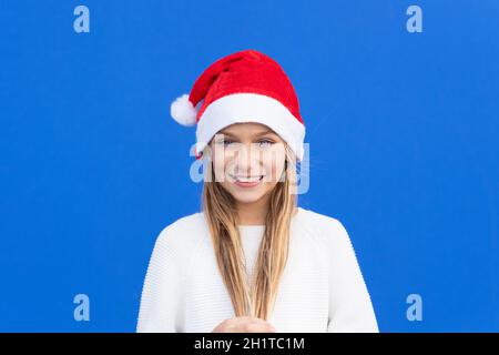 Pleased girl in white sweater and Santa hat holding pigtails smiling away isolated on blue background. High quality photo Stock Photo