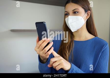 Optimistic girl with FFP2 KN95 mask reads news at smartphone indoors Stock Photo