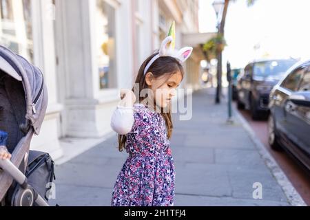 Adorable view of little girl wearing colorful dress and unicorn headband in the street. Portrait of cute young child with unicorn horn and ears standi Stock Photo