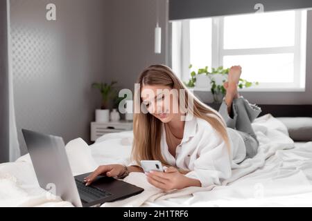 Pretty smiling Caucasian girl lays on bed in bedroom, use notebook and smartphone for online shopping. Online shopping, communication, technology, rem Stock Photo