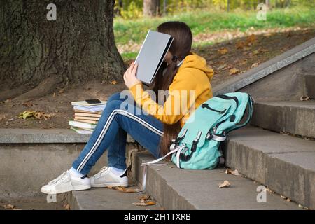 Tired teenage girl reads a book, covers her face with a book sitting on the stairs in the park. Stock Photo