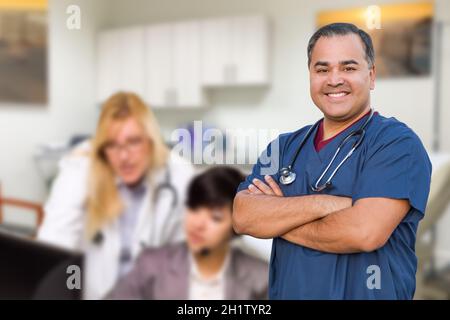 Handsome Hispanic Doctor or Nurse Standing in His Office with Staff Working Behind. Stock Photo