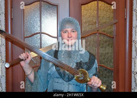 A man dressed up in a knight's hauberk at home in his room. Knight's sword in the hands of a man. Knightly chain armor and sword. Fitting of a knightl Stock Photo