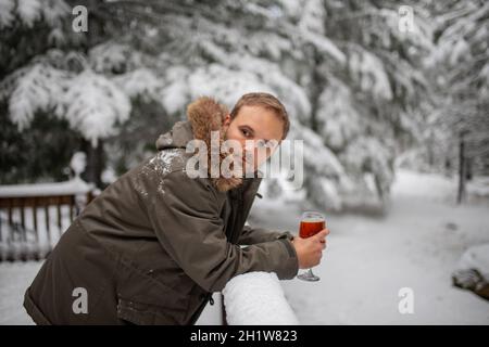 Man holding glass goblet leaning on snow-covered handrail with a snowy forest as background. Thoughtful man resting on wooden balcony and drinking. Re Stock Photo