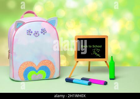 Back to school is written in green letters on a blackboard, colourful pencils and a colourful school backpack stand on a green table over an abstract Stock Photo