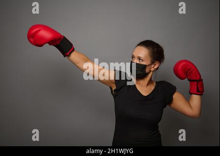 Young athletic woman in safety protective medical mask and red boxing gloves punches an imaginary punching bag isolated over gray background with copy Stock Photo
