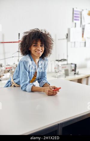 Curly haired African-American lady in denim shirt leans onto cutting table in workshop Stock Photo