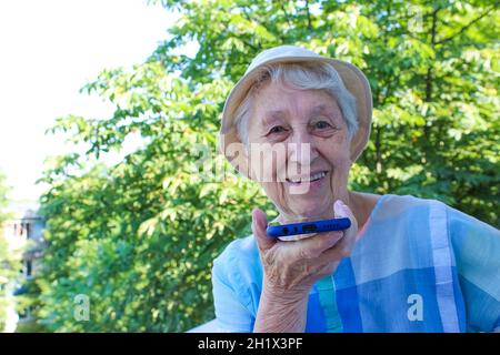 Charming elderly woman in rectangular black rimmed eyewear using voice recognition function on cell phone. Modern senior female holding electronic gad Stock Photo