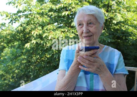 Charming elderly woman in rectangular black rimmed eyewear using voice recognition function on cell phone. Modern senior female holding electronic gad Stock Photo