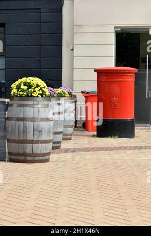 close up of a red mail pillar box with a red recycling bin in the background, outside in a paved street in summer.  Pavement lined with wooden barrels Stock Photo