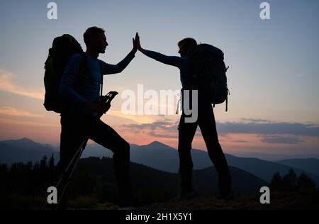 Full length of young man and woman travelers giving high five while hiking together in the mountains during sunset. Concept of travelling, hiking and relationships. Stock Photo