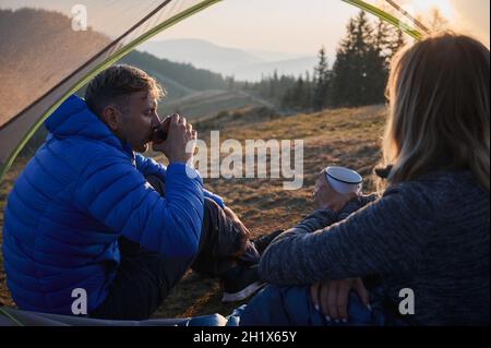 View from inside tent of man and woman sitting inside tent and enjoying pleasant taste of coffee brewed in fresh air. Recreation in tent set up on mountain hill during long hiking. Stock Photo