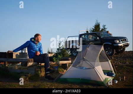 Young man sitting on bench, resting his hand on wooden table located near his tent and looking ahead at the surrounding nature. Black SUV, female silhouette and clear sky on the background. Stock Photo