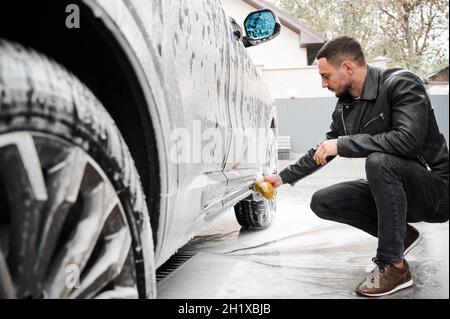 Young man washing car on carwash station outdoor. Handsome driver cleaning automobile, using sponge and soap. Stock Photo