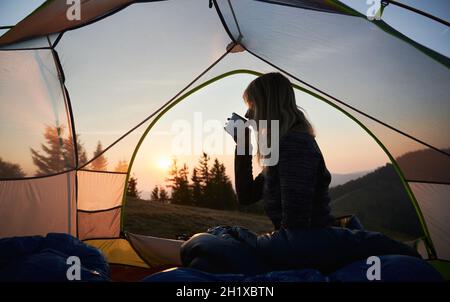 Silhouette of blonde woman drinking hot tea from metal cup, relaxing in her tent and admiring dawn against the backdrop of mountain spaces. Summer camping during sunrise. Stock Photo