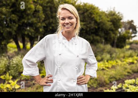 Self-sustainable female chef smiling at the camera cheerfully in an agricultural field. Happy young female chef standing with her arms crossed on an o Stock Photo