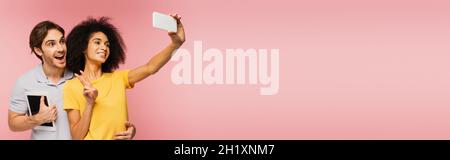 happy latin woman showing victory gesture and taking selfie with amazed man holding digital tablet isolated on pink, banner Stock Photo