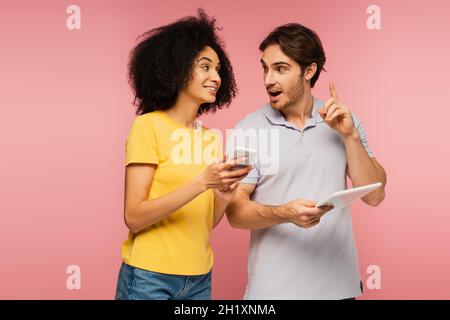 amazed man with digital tablet showing idea gesture near smiling hispanic woman with  cellphone isolated on pink Stock Photo