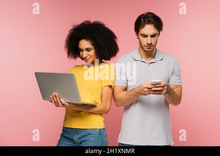 smiling hispanic woman using laptop near serious man typing on mobile phone isolated on pink Stock Photo