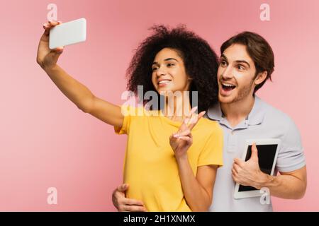amazed man with digital tablet hugging hispanic girlfriend showing victory sign while taking selfie isolated on pink Stock Photo