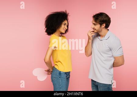 cheerful hispanic woman holding paper heart behind back near curious boyfriend isolated on pink Stock Photo