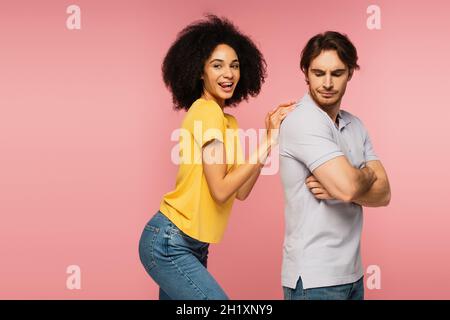 cheerful latin woman sticking out tongue while touching back of upset man standing with crossed arms isolated on pink Stock Photo