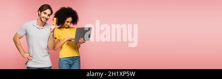 happy man showing credit card near hispanic girlfriend using laptop isolated on pink, banner Stock Photo
