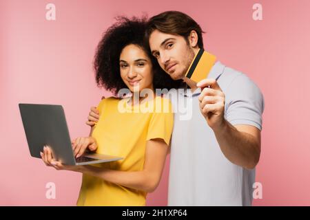 man showing credit card and embracing hispanic girlfriend using laptop isolated on pink Stock Photo