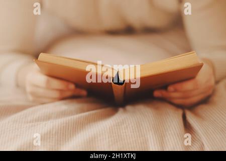 A man in a beige sweater is lying on a soft bed with a striped blanket and reading an old interesting hardcover book. Recreation and entertainment. Ho Stock Photo