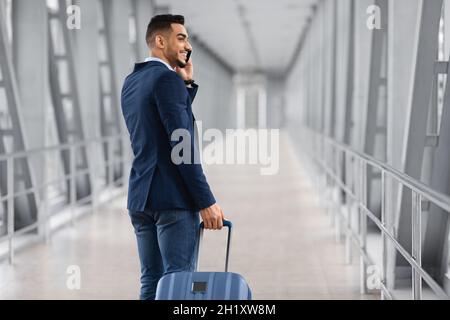 After Arrival. Smiling Arab Businessman Talking On Cellphone While Passing Airport Gate Stock Photo