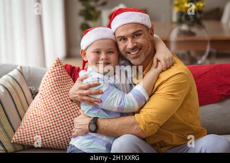 Portrait of caucasian father and son hugging and smiling at home during christmas Stock Photo