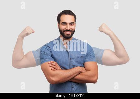 Successful positive bearded man in blue shirt folding hands on chest and looking at camera with toothy smile showing strong big arms in shadow. Indoor studio shot isolated on gray background Stock Photo