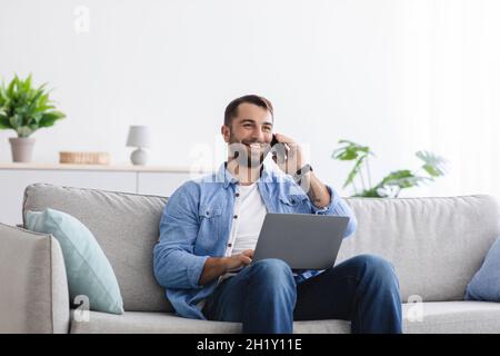 Cheerful handsome mature caucasian guy with beard talking by phone with client and working on laptop Stock Photo