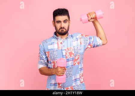 Self confident young adult bearded man wearing casual style blue shirt holding dumbbells, pumping muscles, having funny facial expression. Indoor studio shot isolated on pink background. Stock Photo