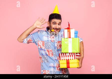 Funny bearded attractive man wearing casual blue shirt and cone, holding horn in mouth, waving hand to camera, celebrating birthday, rejoicing. Indoor studio shot isolated on pink background.
