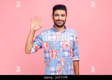 Hello, welcome. Portrait of happy young adult dark haired bearded man in blue shirt raising palm to wave hi, greeting with hospitable friendly smile Indoor studio shot isolated on pink background. Stock Photo