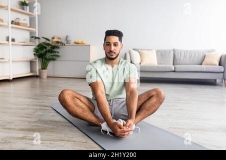 Home workout. Sporty young Arab guy stretching on mat in living room, practicing yoga, full length Stock Photo