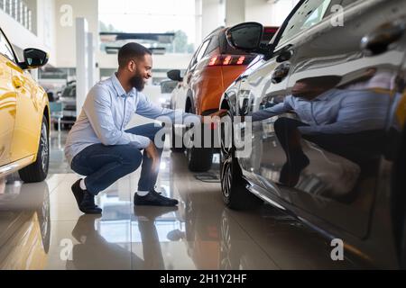 Black Male Customer Choosing New Automobile In Dealership Center, Examining Wheel Rims Stock Photo