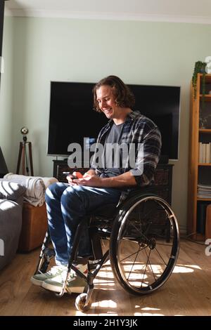 Caucasian disabled man sitting on wheelchair smiling while using smartphone at home Stock Photo