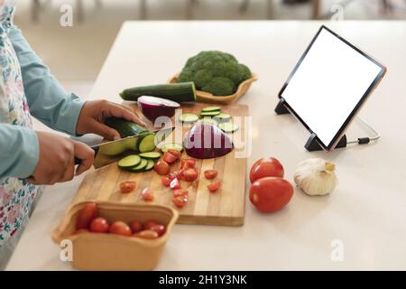 Hands of african american woman using tablet with copy space and cutting vegetables in kitchen Stock Photo