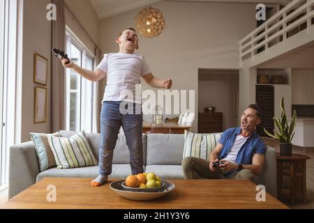 Caucasian boy celebrating standing on the table while playing video games with his father at home