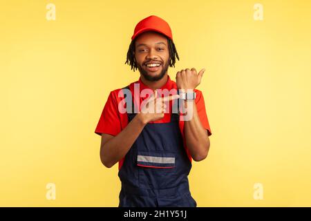 We do on time. Smiling happy courier or mechanic standing pointing at wristwatch, showing work time, bearded worker wearing overalls. Indoor studio shot isolated on yellow background. Stock Photo