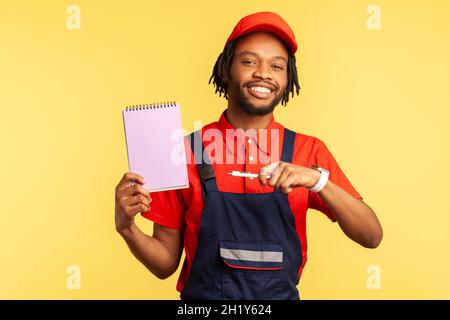 Portrait of workman wearing blue overalls and red cap, holding pointing at paper notebook with pleasant smile, presenting copy space. Indoor studio shot isolated on yellow background. Stock Photo