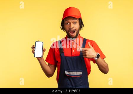 Shocked workman in overalls pointing at mobile phone with blank screen, surprised by online delivery order app, house repair maintenance services. Indoor studio shot isolated on yellow background. Stock Photo
