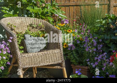 Overgrown wicker chair in small garden with stone plaque reading 'One who plants a garden plants happiness' Stock Photo