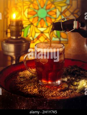 Beer in a pint glass with handle poured from a traditional flip top bottle on beer barrel with hops & barley lit oil lamp and stained glass in back Stock Photo