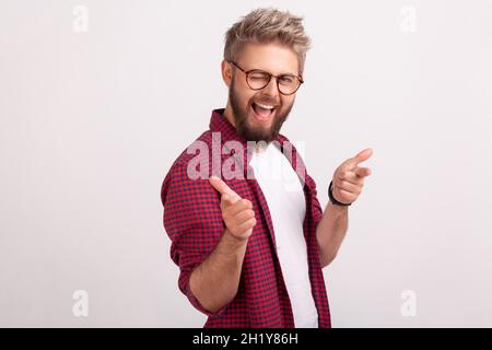 Portrait of pleased bearded male in eyeglasses smiling and winking on camera, gesturing hey you sign. Indoor studio shot isolated on gray background Stock Photo