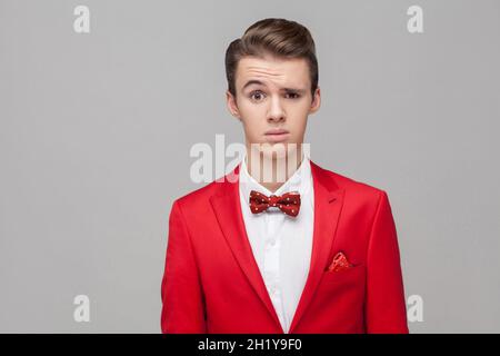 Portrait of confused young man with stylish hairdo in red tuxedo and bow tie looking with surprised and puzzled face, thinking what is going on. indoor studio shot isolated on gray background Stock Photo