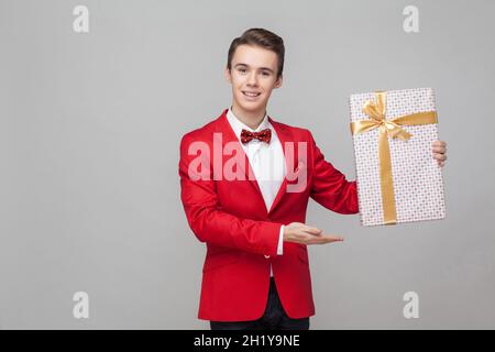 Portrait of cheerful elegant handsome gentleman with stylish hairdo in red tuxedo and bow tie pointing at big wrapped present box, showing holiday gift. indoor studio shot isolated on white background Stock Photo